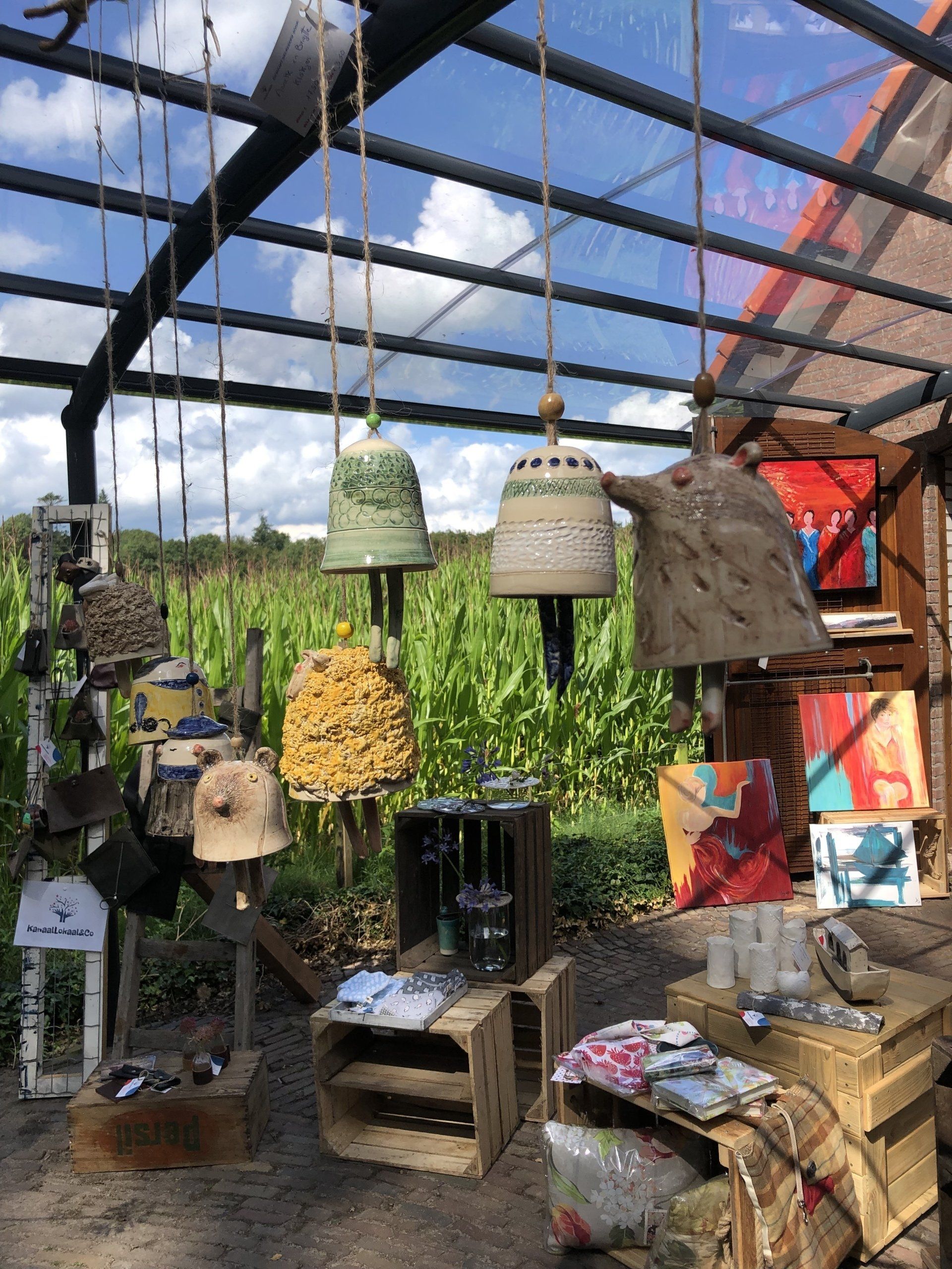 Binnen in een constructie met houten kisten en vitrines hangen bellen; buiten is een maïsveld zichtbaar.