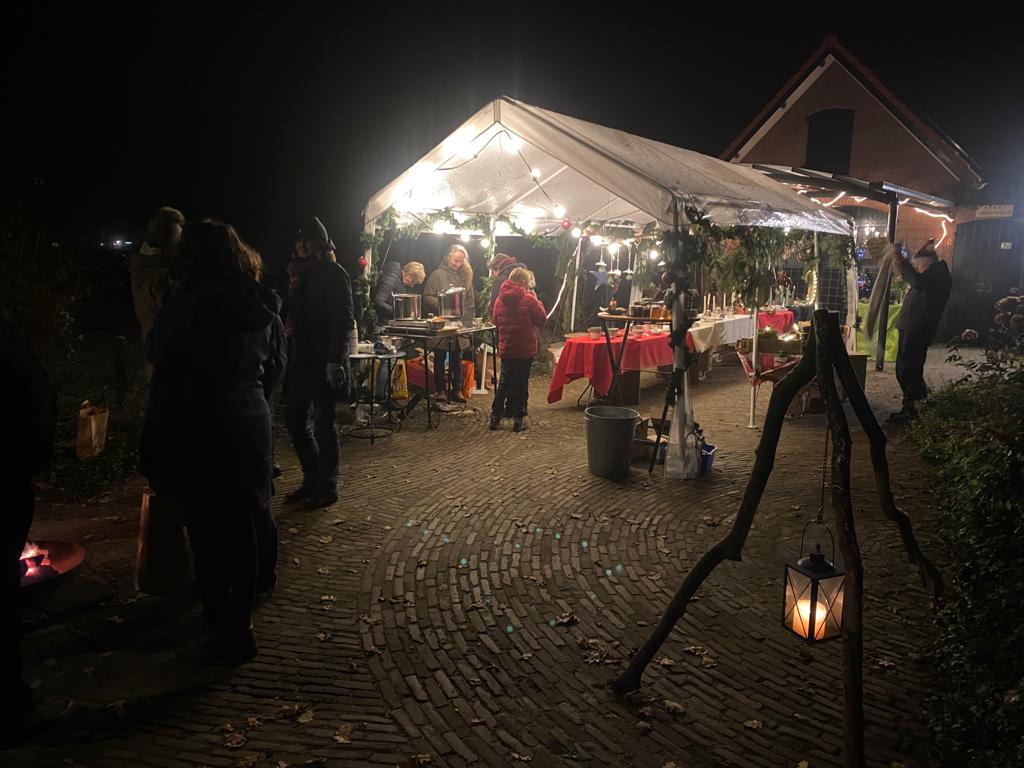 Nachtscène: Mensen op een openluchtmarkt onder een verlichte tent. Een lantaarn hangt aan een houten statief.