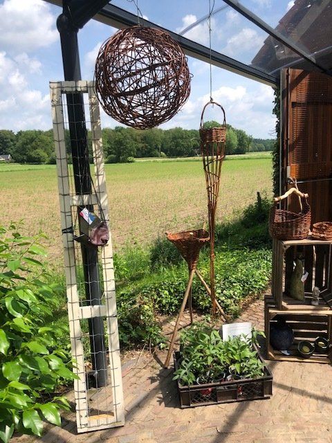 Een kasinterieur met hangende decoraties en planten, met uitzicht op een groen veld onder een zonnige hemel.