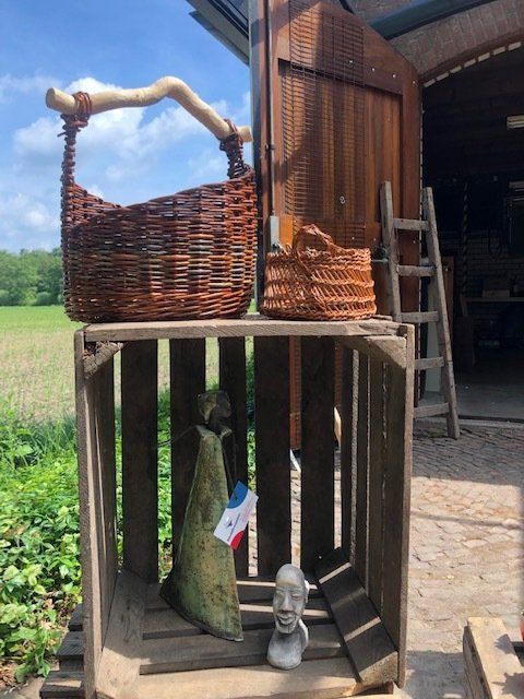 Rieten manden en een sculptuur staan uitgestald op een houten kist in de buitenlucht. Op de achtergrond zijn een veld en een houten deur te zien.