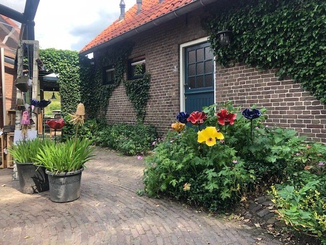 Bakstenen gebouw met een rood pannendak en een blauwe deur. Kleurrijke bloemen bloeien ervoor en groene klimop klimt tegen de muur omhoog.