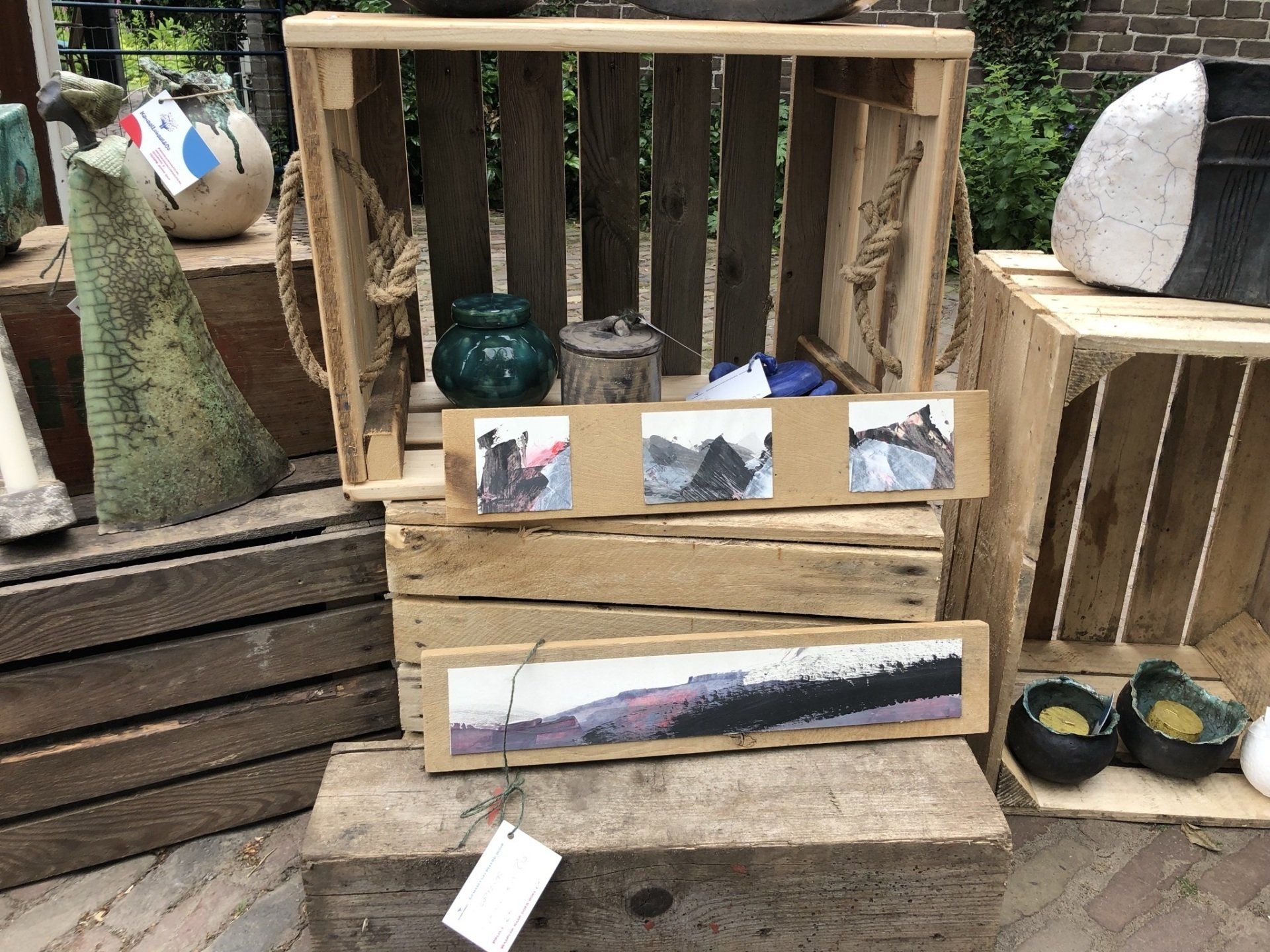 Aardewerk en kunstwerken tentoongesteld op houten kisten; op de voorgrond bevindt zich een landschapsschilderij.