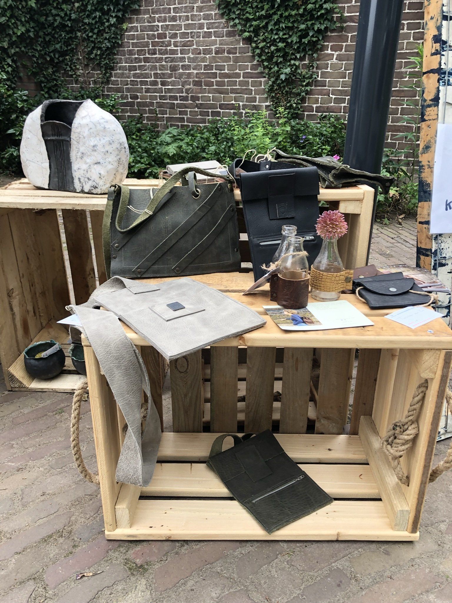 Handgemaakte tassen en accessoires tentoongesteld op houten kratten in de buitenlucht.