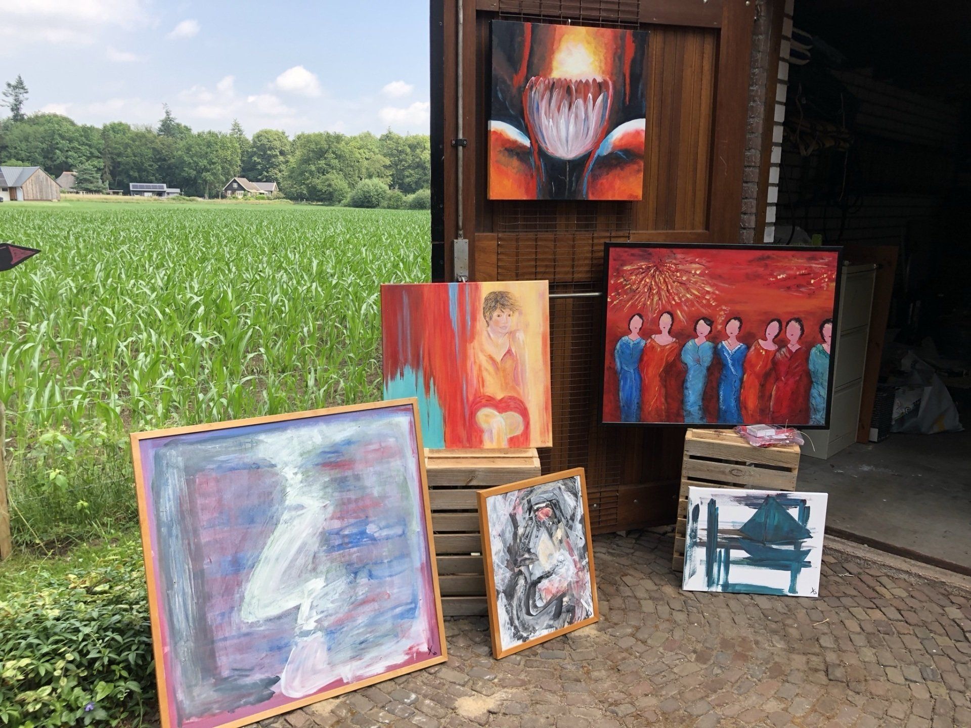 Schilderijen tentoongesteld in de buitenlucht tegen een rustieke houten constructie, met een veld op de achtergrond.