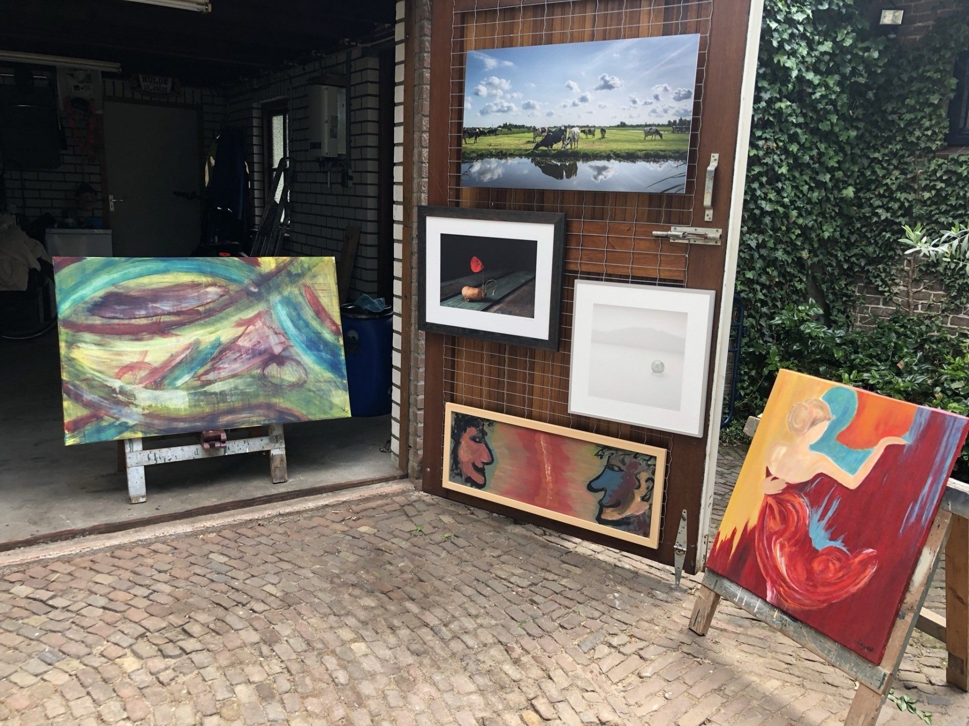 Kunstwerken tentoongesteld: schilderijen op schildersezels, gemonteerd aan een houten wand, buiten.