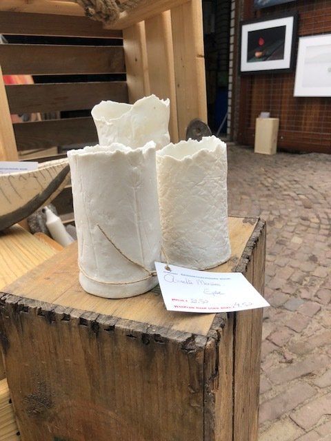Drie witte keramische vazen met bladmotieven staan op een houten ondergrond, met een visitekaartje ernaast.