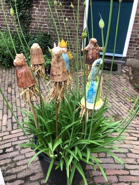 Keramische figuren met rokjes en kroontjes van stro, in een pot met groene planten.