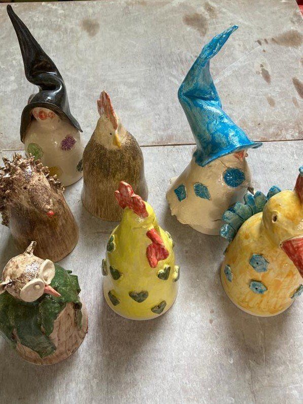 Keramische kippensculpturen met diverse hoeden en versieringen, op een lichtgekleurde ondergrond.