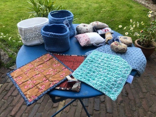 Handgemaakte gehaakte en gequilte artikelen op een blauwe tafel buiten. Inclusief manden, pannenlappen en kleine kussens.