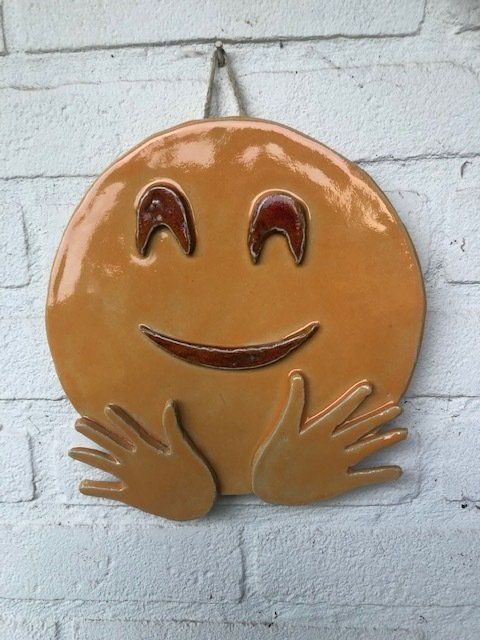 Een lachende, keramische emoji met armen, bruine ogen en mond, hangt aan een witte bakstenen muur.