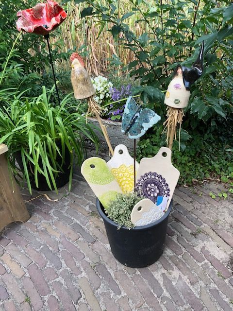 Zwarte emmer met keramische tuindecoraties, waaronder bloemen, vlinders en snijplankvormen. Buitenomgeving met planten en bakstenen muren.