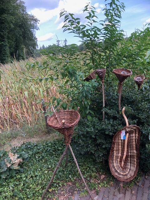 Rotansculpturen in een tuinomgeving, naast hoog gras en weelderige struiken.