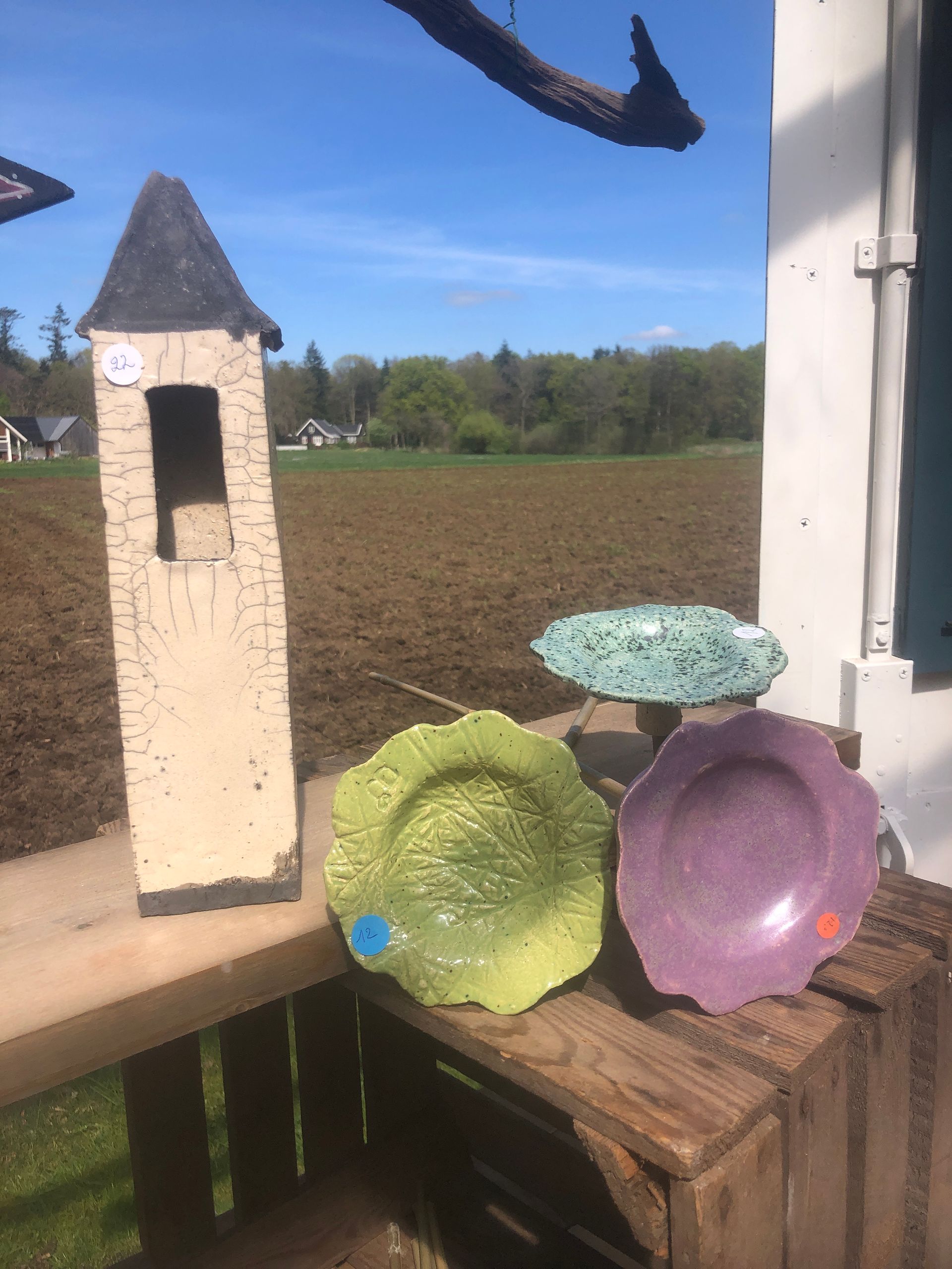 Keramische toren en kleurrijke borden tentoongesteld op een houten standaard in de buitenlucht, met een veld op de achtergrond.