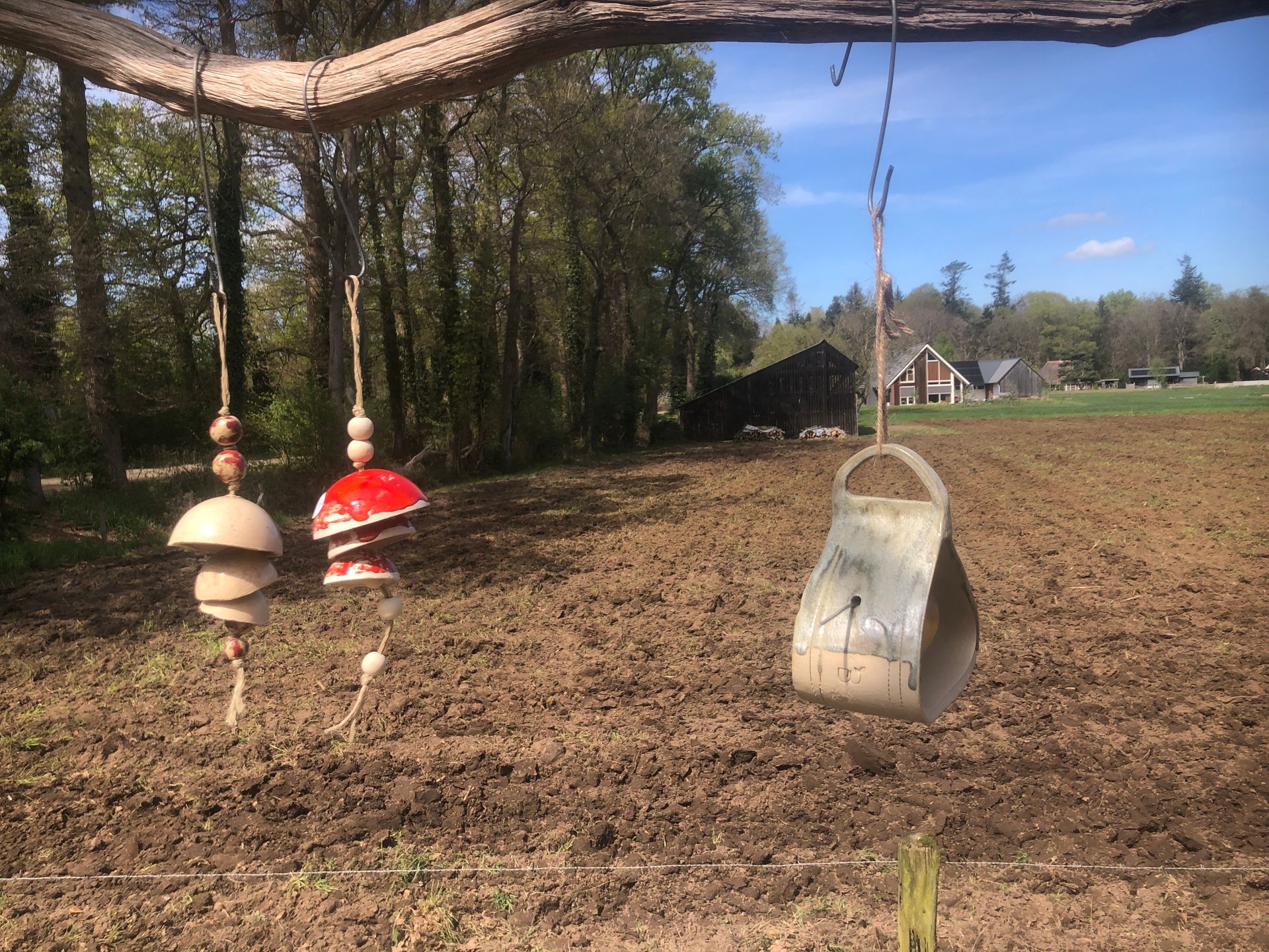 Een windgong en een bel hangen in een tuin met op de achtergrond een geploegd veld, bomen en gebouwen.