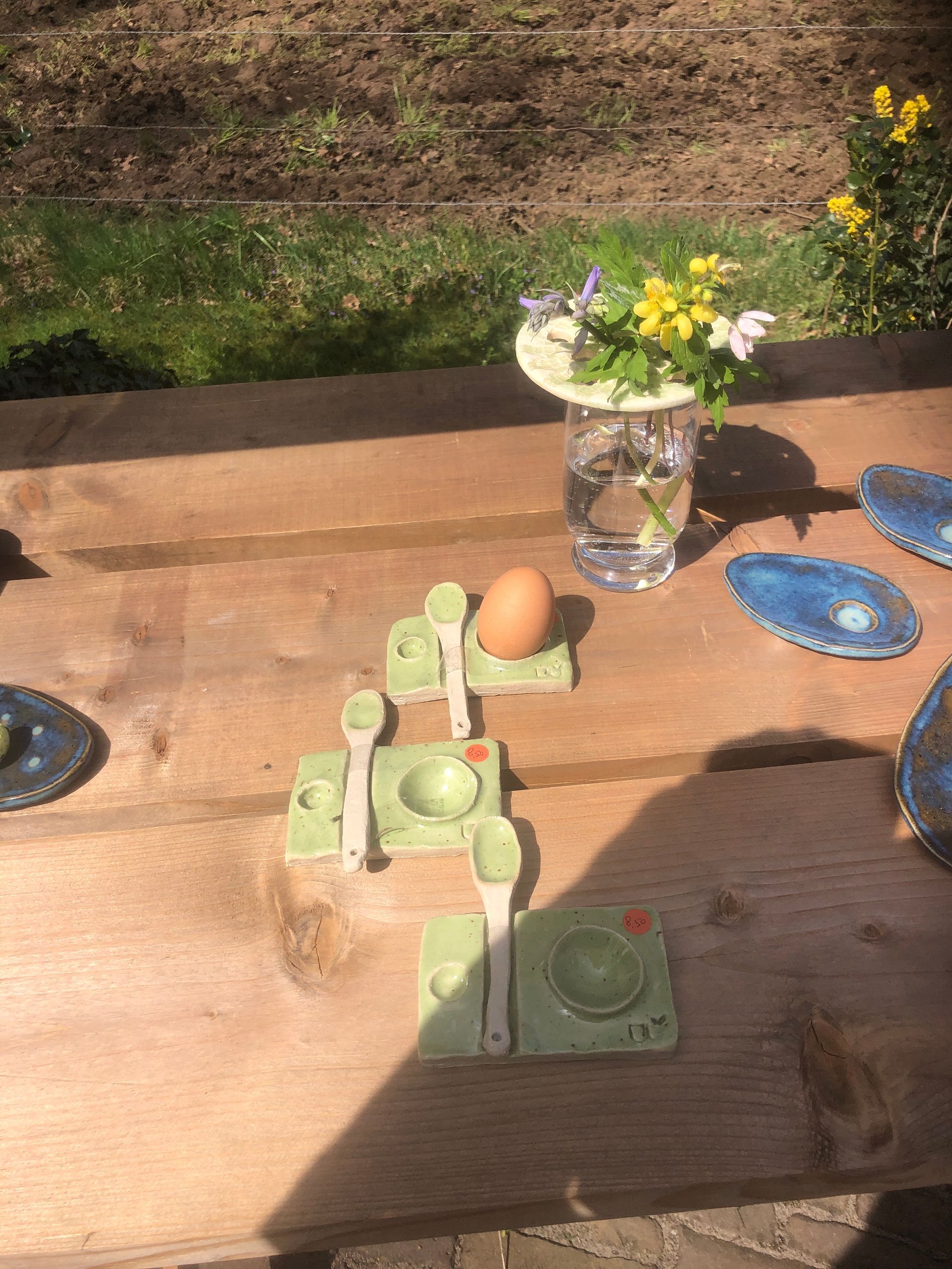 Houten tafel met keramische eierhouders, lepels, een ei en een glas met bloemen.