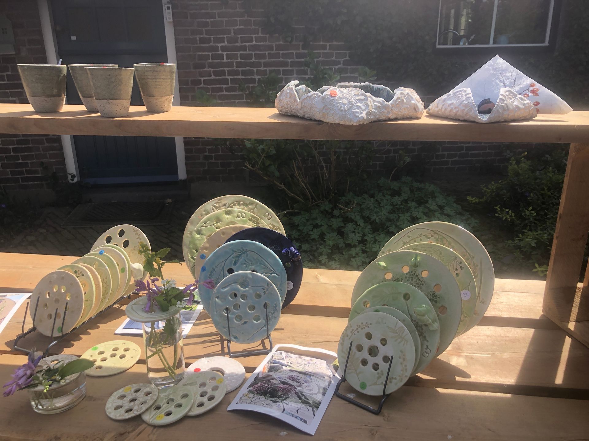 Handgemaakt aardewerk tentoongesteld op een houten tafel buiten. Met kopjes, kommen en borden in neutrale tinten en groen.