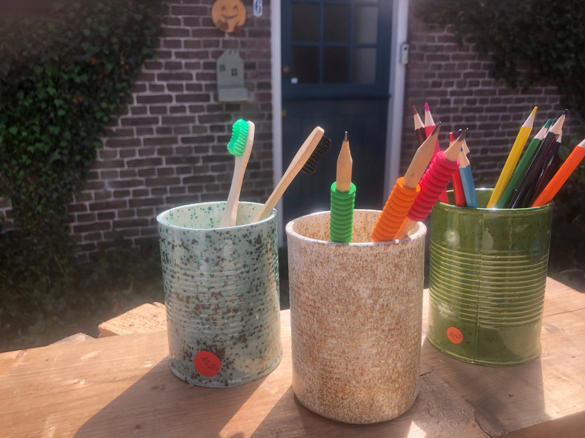 Drie versierde blikken, met daarin een tandenborstel en potloden, staan op een houten ondergrond, met op de achtergrond een bakstenen muur en een deur.