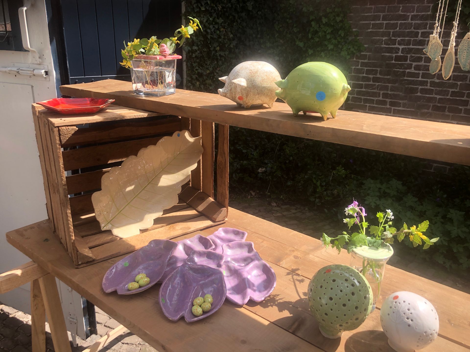 Houten display met keramische kunst: schalen, sculpturen en kleine plantjes in een zonnige buitenomgeving.