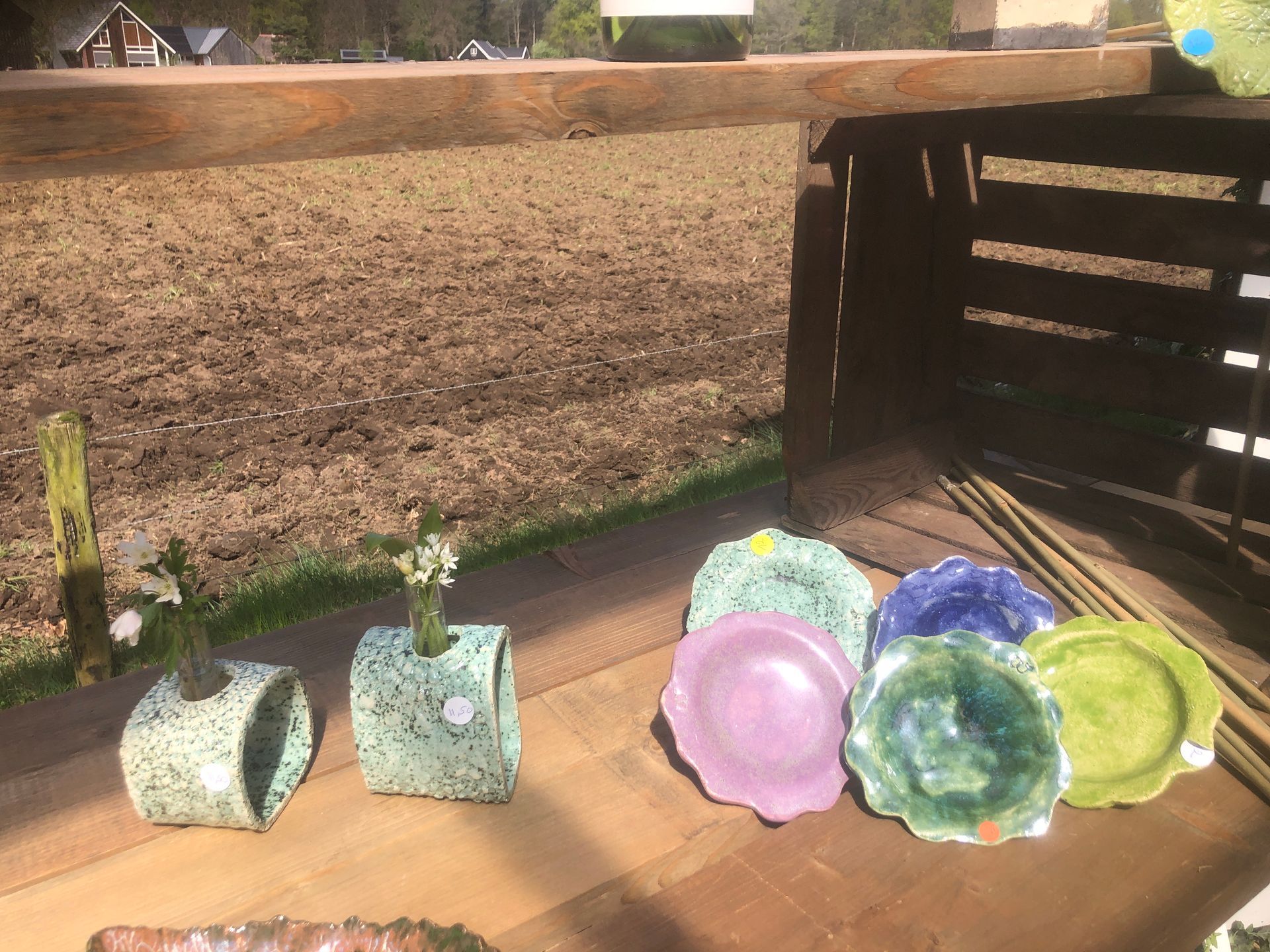 Keramische potten en borden in diverse kleuren staan op een houten ondergrond, tegen een bruine veldachtergrond.