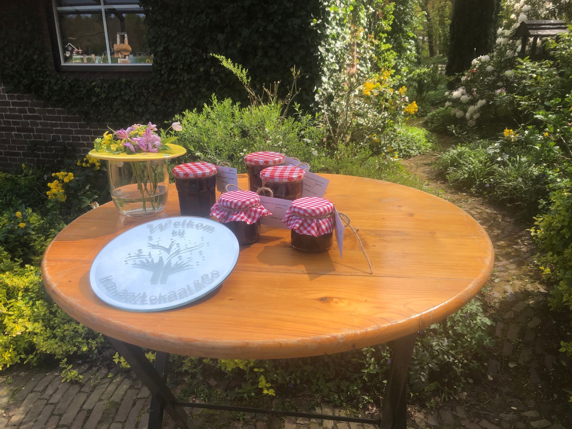 Ronde houten tafel buiten met potten jam, een bord en bloemen in een tuinomgeving.