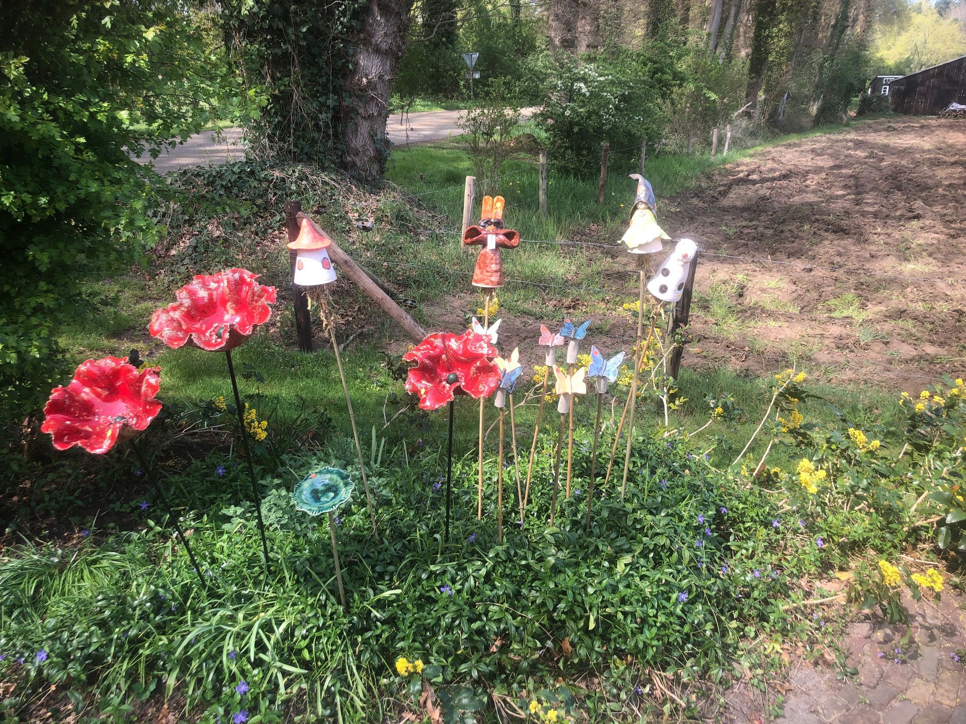 Een tuin met glazen bloemsculpturen en speelse versieringen. Groen gebladerte op de voorgrond, bomen en een zandpad op de achtergrond.