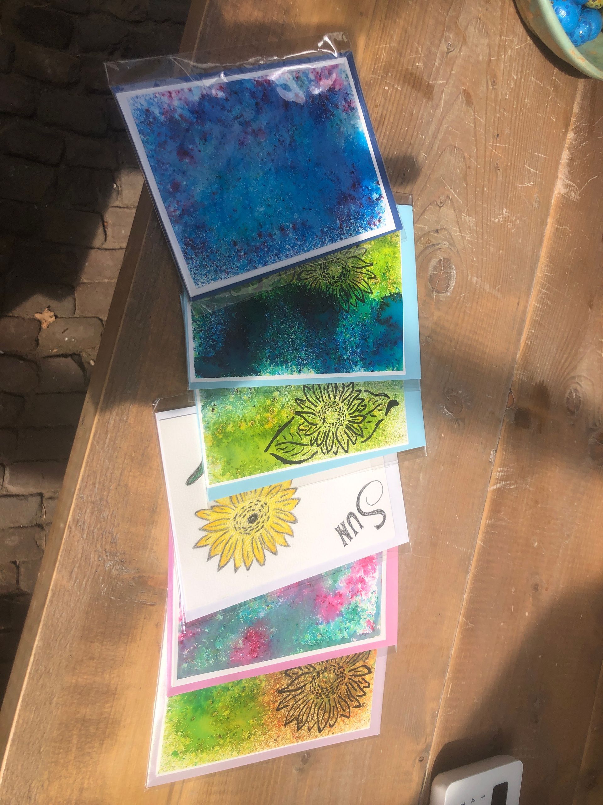 Een stapel kleurrijke, met glitter gevulde kunstkaarten op een houten ondergrond. De ontwerpen omvatten bloemen, woorden en abstracte patronen.