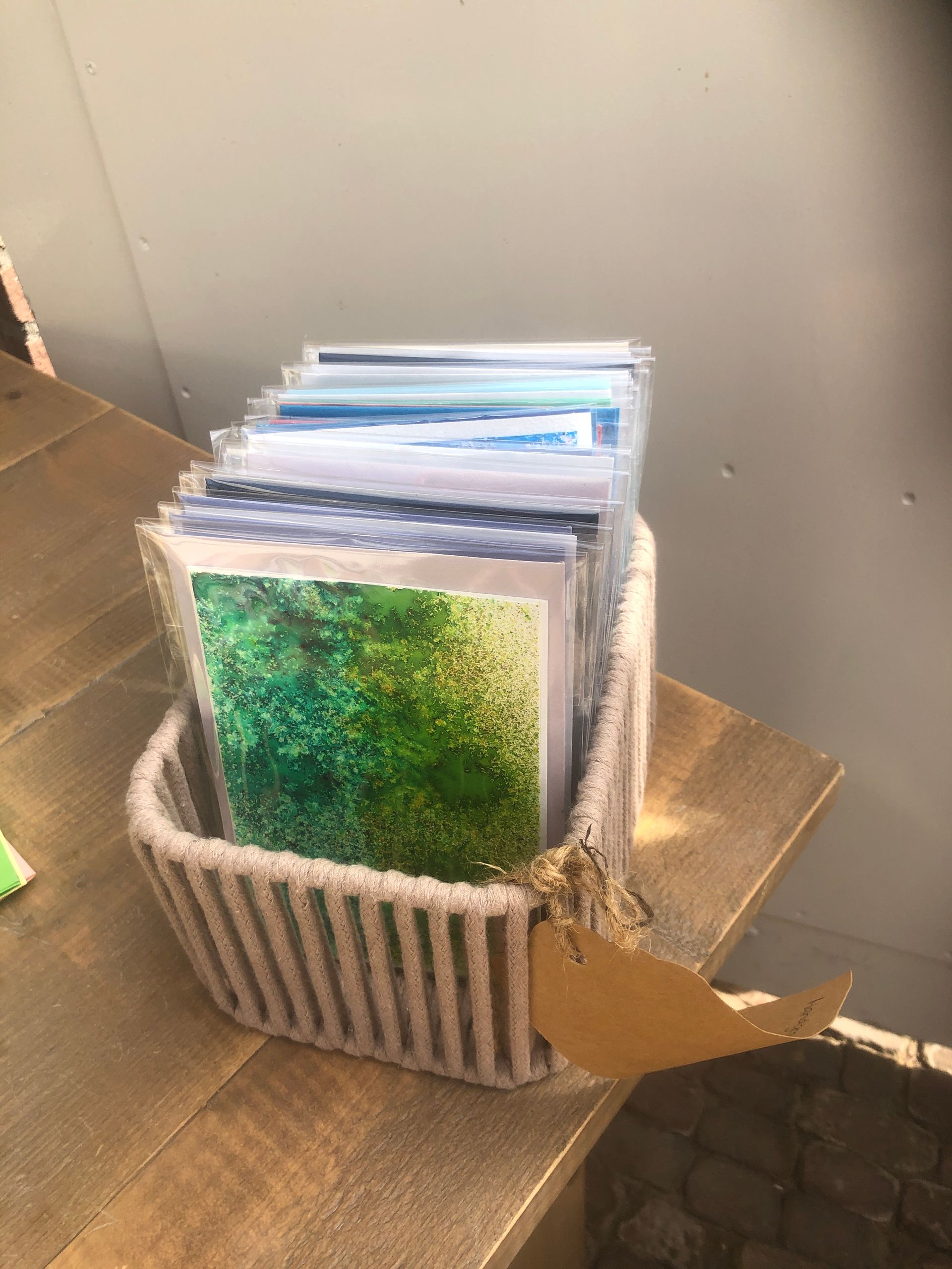 Een gevlochten mand met daarop een stapel wenskaarten met groene en blauwe afbeeldingen ligt op een houten ondergrond.