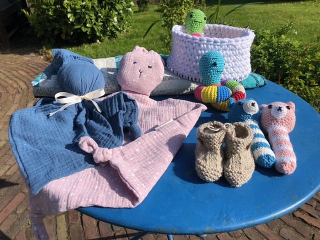 Handgemaakte babyartikelen: roze en blauwe stof, speelgoed, gehaakte babyschoentjes en een mandje op een blauwe tafel buiten.
