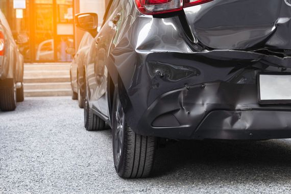 A black car with a damaged rear bumper parked on a gravel lot in front of a building.