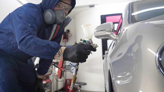 A worker in a protective suit and respirator uses a spray gun to paint the side of a silver car in a workshop.