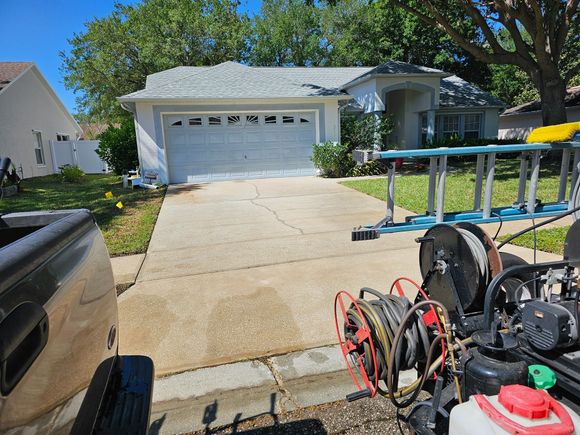 A pressure washing setup in front of a house with a concrete driveway and a truck parked to the left.