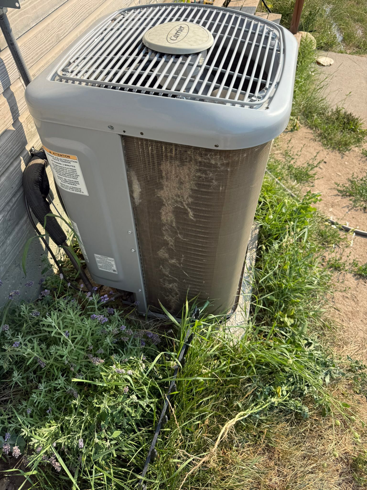 Outdoor air conditioner unit with a damaged side panel, standing in overgrown grass by a fence.