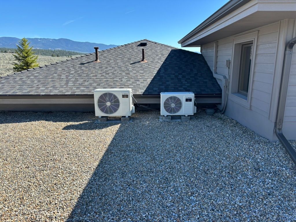 Two white outdoor AC units on a gravel roof beside a house, with mountains in the background
