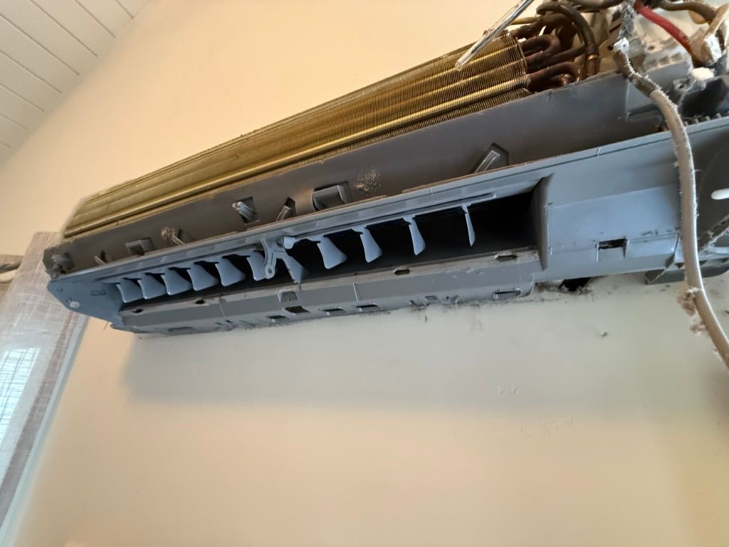 Broken wall-mounted air conditioner with open vents and exposed wiring near a window