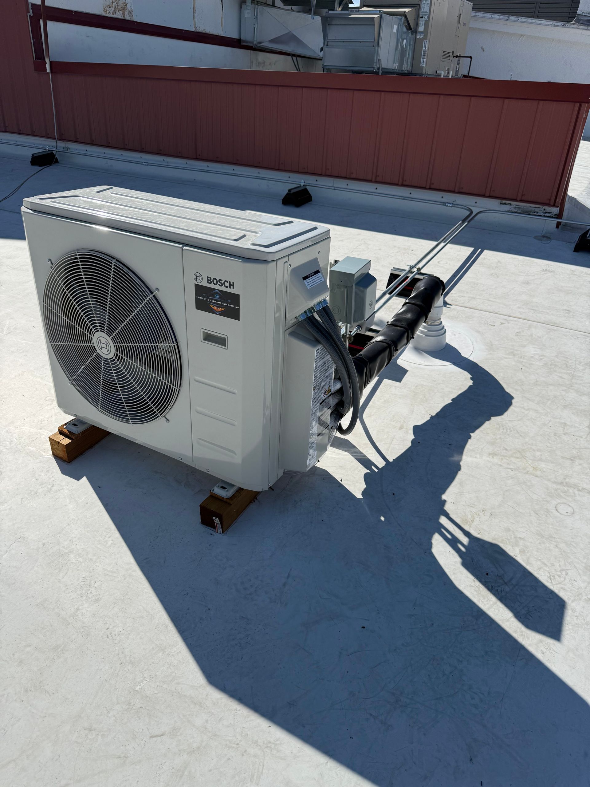 Outdoor air conditioner condenser unit on a rooftop, with exposed copper pipes and sunlight casting shadows.