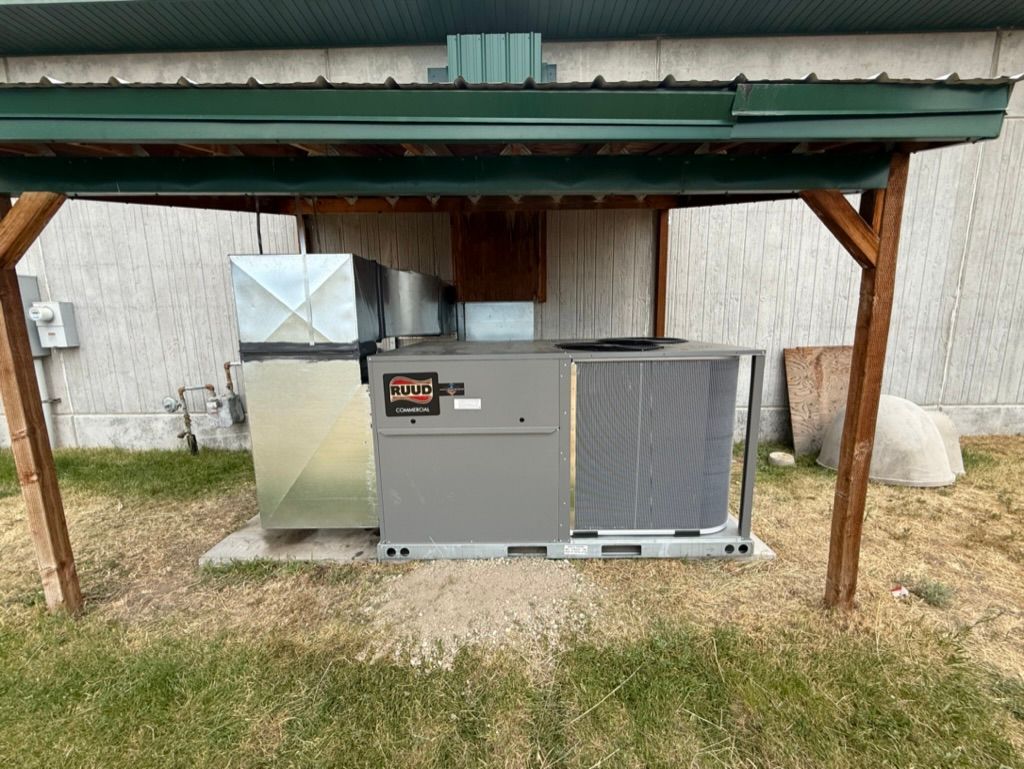 Outdoor HVAC unit under a green-roofed shelter beside a building, with a metal duct on the left.