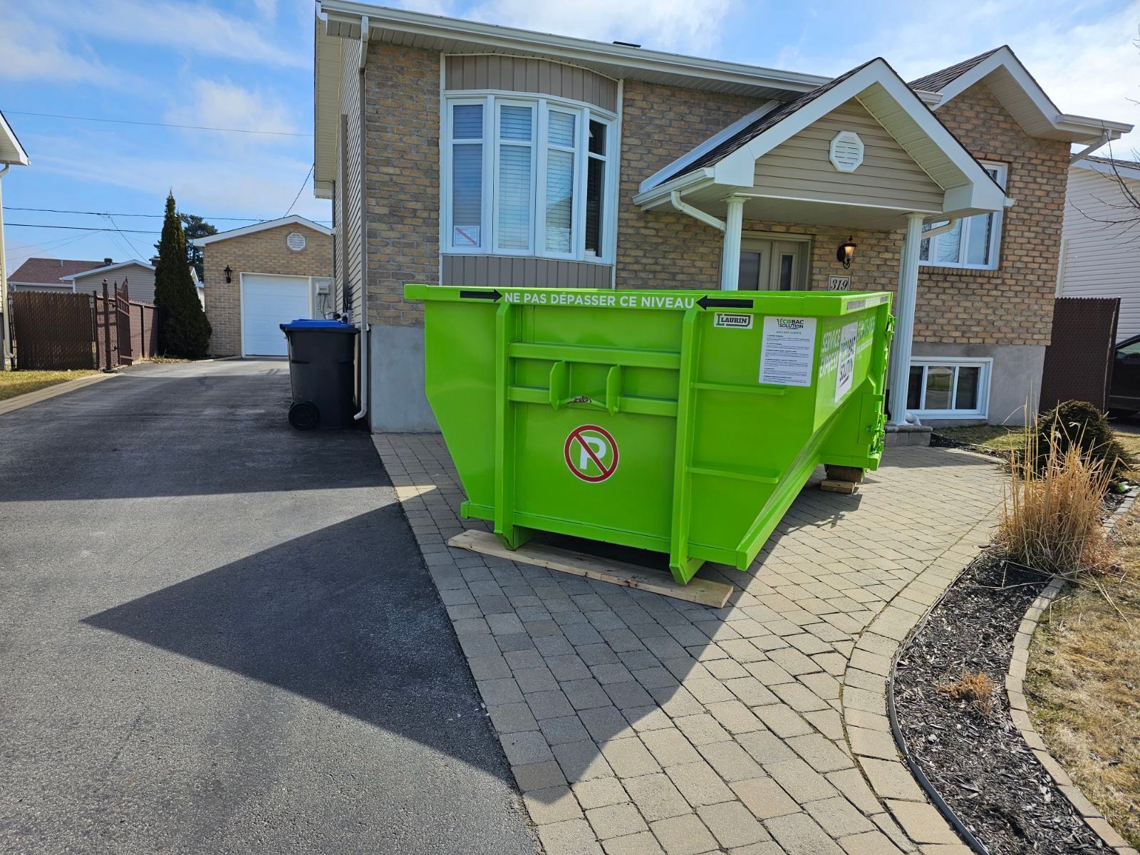 Une grande benne à ordures en métal vert vif est posée sur une allée pavée devant une maison en briques.