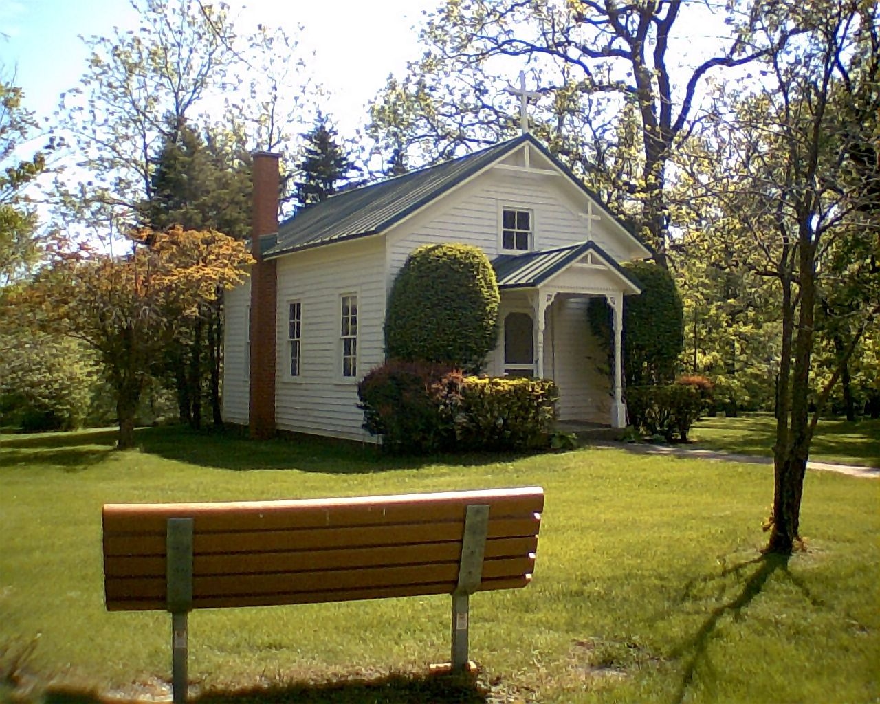A small, white wooden chapel with a brick chimney and a front porch sits in a green grassy area with a wooden park bench.