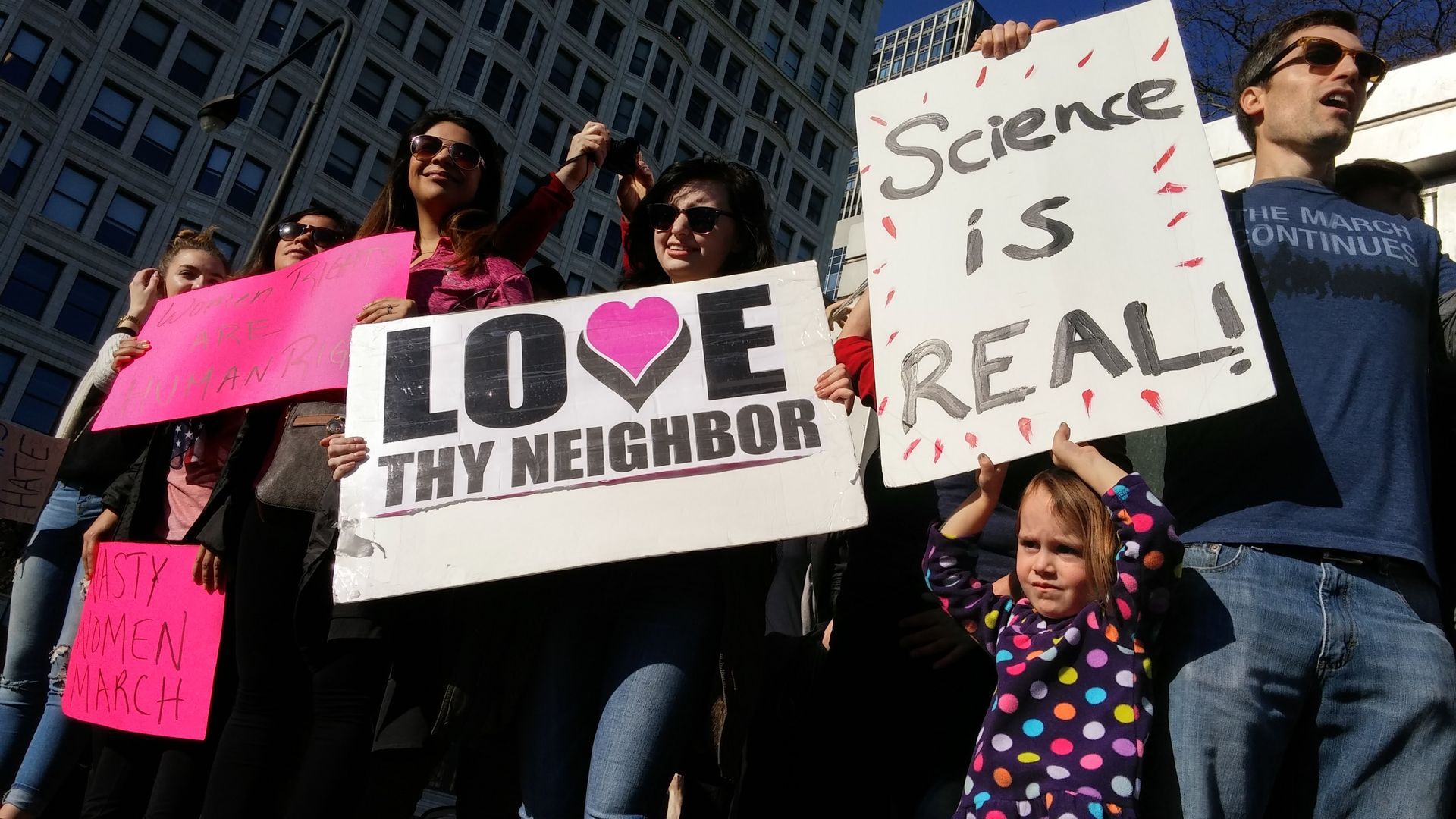 A crowd of people stands at a city intersection, one person holding a sign that says,