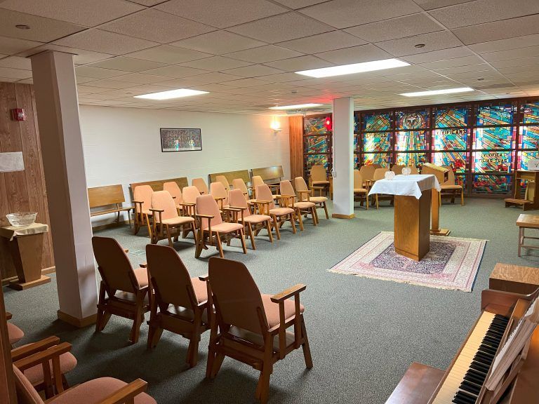 A small chapel interior with rows of wooden chairs, a podium with a white cloth, and a stained-glass window background.