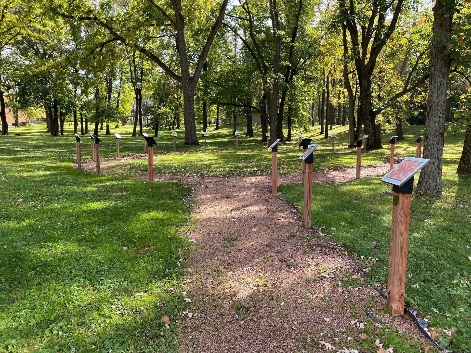 A gravel walking path winds through a wooded park, featuring several upright wooden signs with information plaques.
