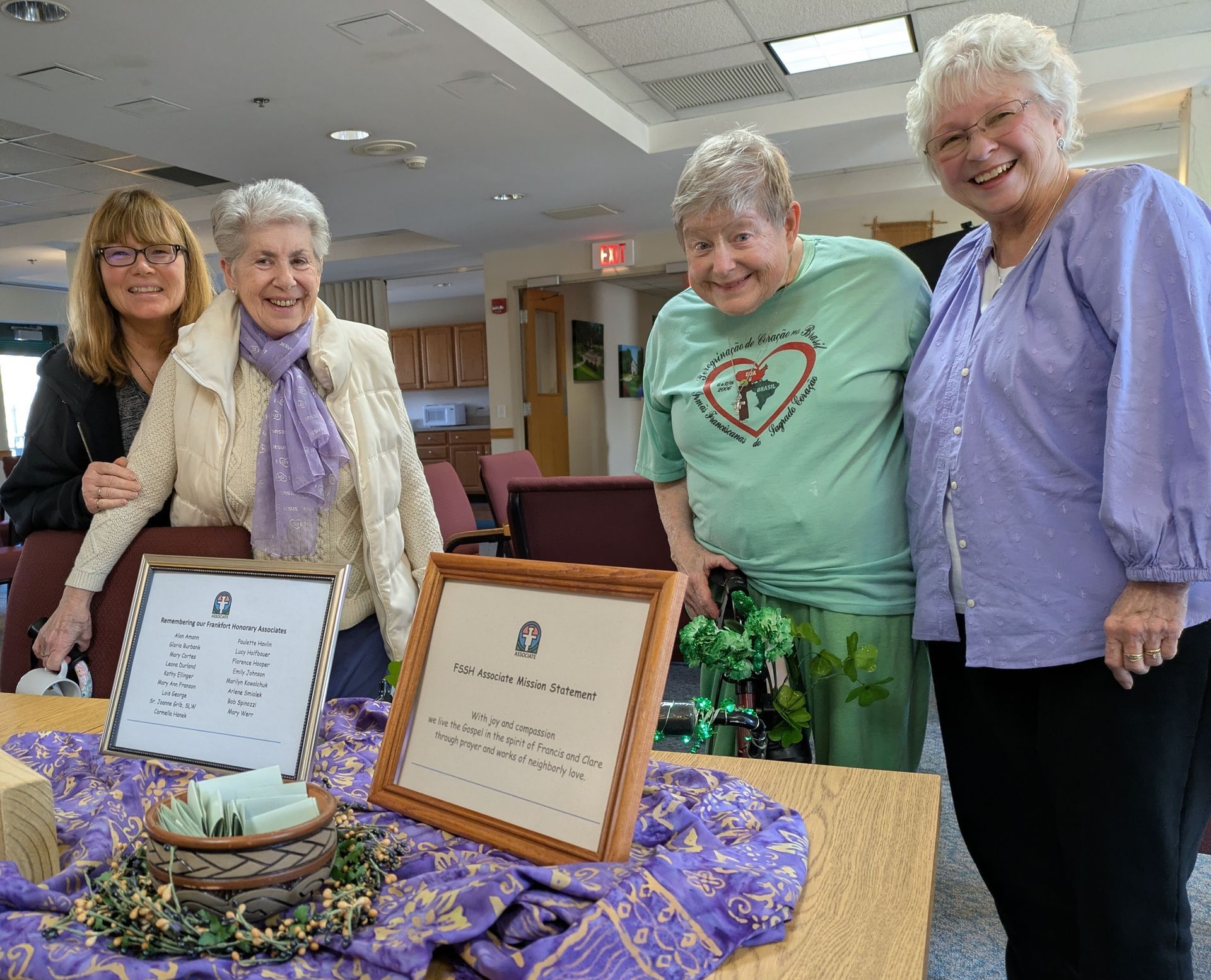 Four people smiling behind a table with two framed signs and a small bowl, set in a bright community room.