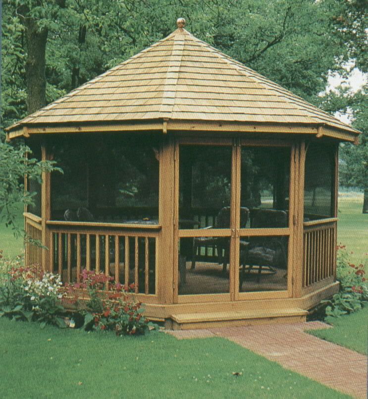 A wooden gazebo with a shingled roof, screened walls, and railing, surrounded by grass and flowers in a park setting.