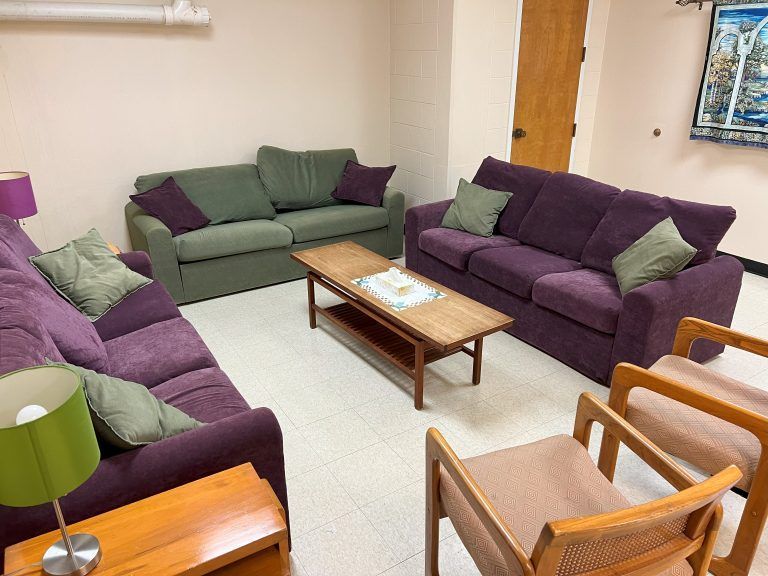 A lounge with a green sofa, two purple sofas, a wooden coffee table, and two wooden chairs arranged on a light floor.