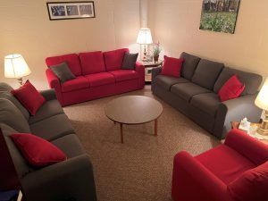 A cozy living room with a red sofa, a grey sofa, a matching armchair, a round coffee table, and lamps on a beige carpet.