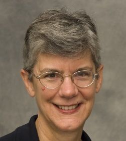 A person with short, grey hair and glasses smiling against a plain, neutral grey background.