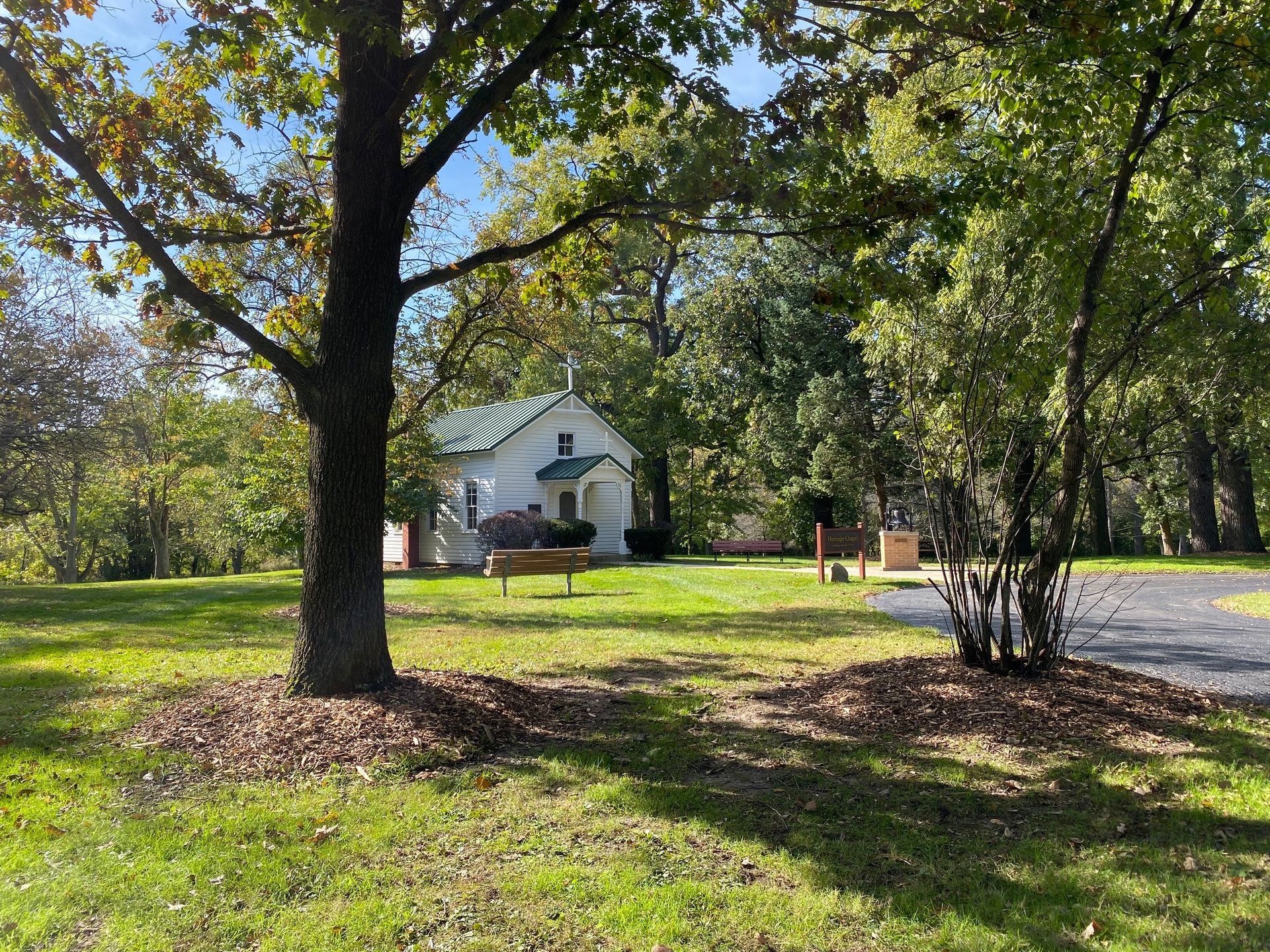 A small white cottage sits among mature trees in a grassy park setting, with a paved path visible on the right.