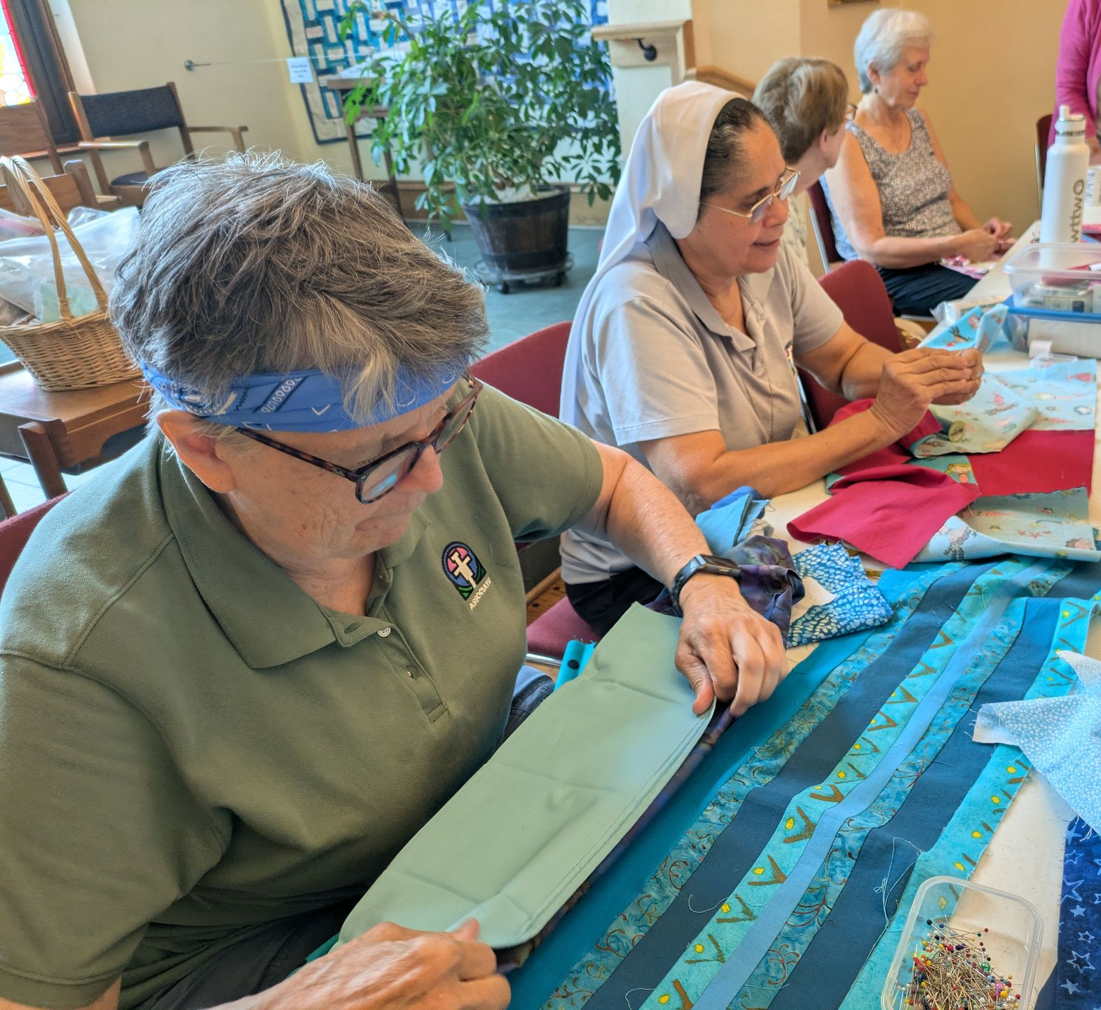 Three people sit at a long table indoors, focused on sewing or crafting with various blue and green fabric scraps.