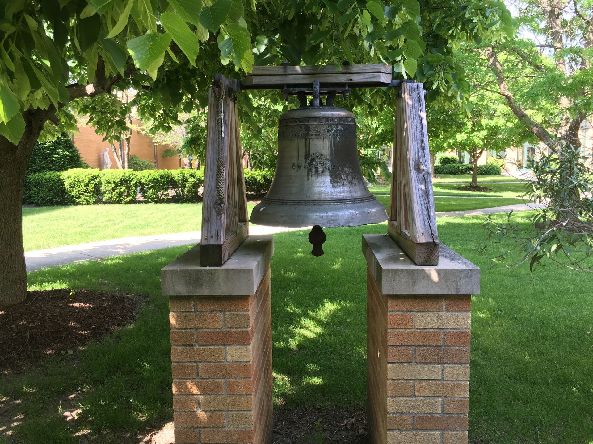 A large metal bell hangs from a wooden frame supported by two brick pillars in a grassy park setting.