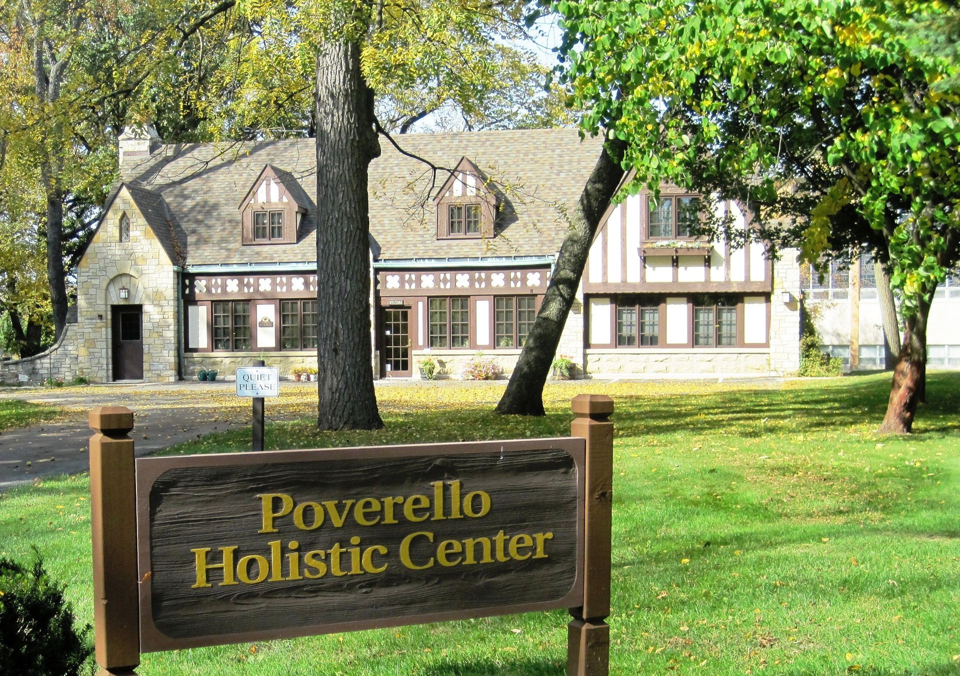 The Poverello Holistic Center, a stone and timber-framed building, sits on a grassy lawn with a prominent wooden sign.